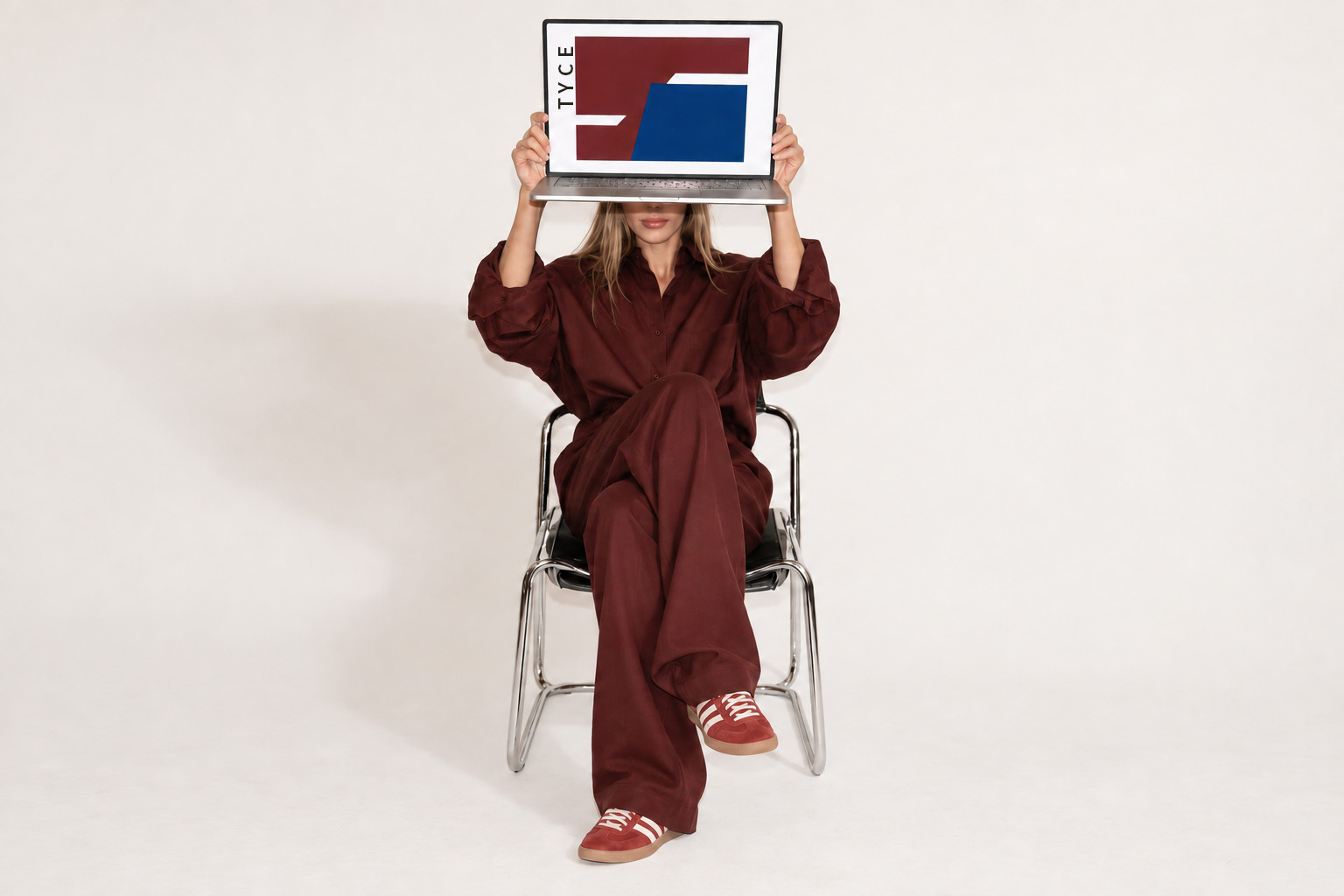Seated woman in burgundy holding a laptop over her face; the screen shows TYCE artwork in burgundy and cobalt